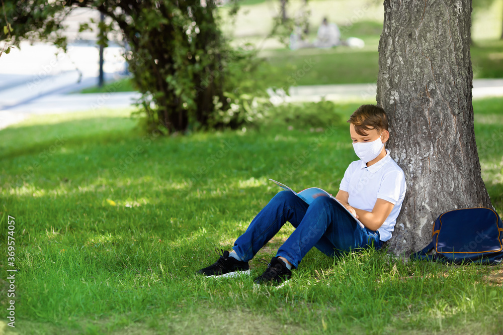 One boy wearing mask is sitting on the grass at park, reading book, doing home work after school during covid-19 pandemic time. New normal of coronavirus epidemic and education concept One boy wearing mask is sitting on the grass at park, reading book, doing home work after school during covid-19 pandemic time. New normal of coronavirus epidemic and education concept