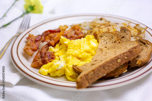 A view of a breakfast plate, featuring bacon, scrambled eggs, wheat toast, and shredded hash browns, in a restaurant or kitchen setting.