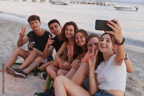 Group of friends taking selfie with mobile phone sitting on the beach. Teenagers have fun on the beach during summer holidays, laughing and taking pictures of themselfs.