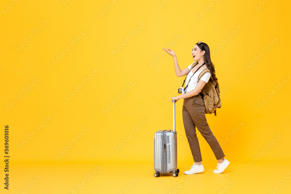 Full length portrait of beautiful smiling young Asian woman tourist with trolley bag looking and opening hand to copy space isolated on colorful yellow studio background Full length portrait of beautiful smiling young Asian woman tourist with trolley bag looking and opening hand to copy space isolated on colorful yellow studio background
