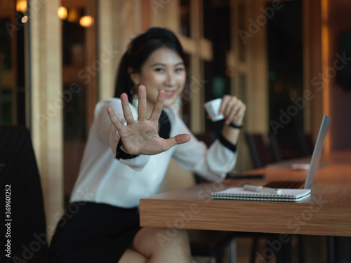 Portrait of cheerful businesswoman sitting in co working space with raised palm and smiling