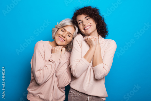 Delightful caucasian women dreaming about something and smile on a blue studio wall posing close to each other