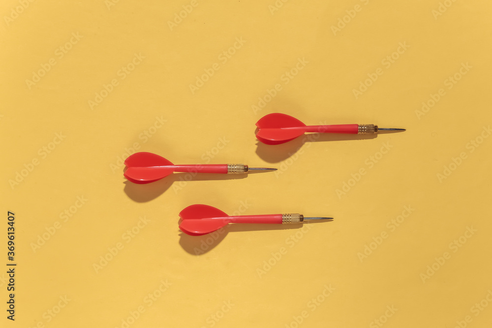 Three red plastic darts with metal tip on yellow bright background with deep shadow. Top view. Flat lay Three red plastic darts with metal tip on yellow bright background with deep shadow. Top view. Flat lay