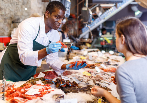 Positive male African American seller serving customer fresh shrimps at seafood market