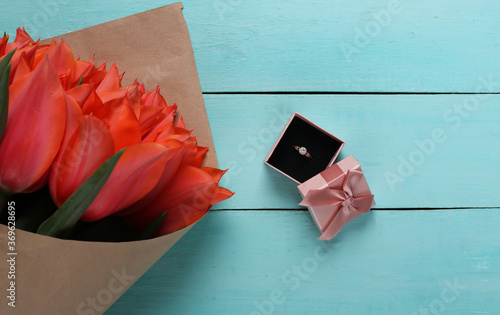 Ring with a diamond in a gift box and a bouquet of red tulips on blue wooden background. Birthday, Valentine's Day or marriage proposal. Top view photo