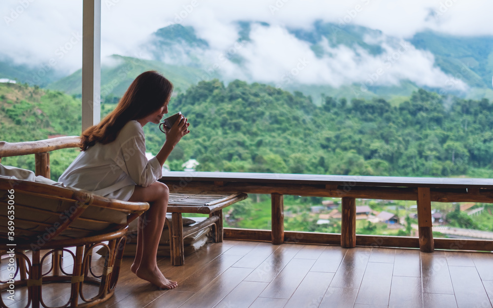 Portrait image of a beautiful asian woman holding and drinking hot coffee , sitting on balcony and looking at mountains and green nature