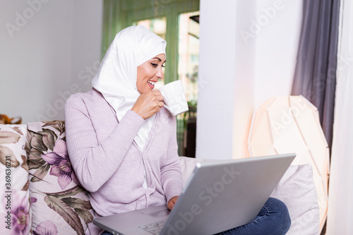 Happy pretty Muslim woman using laptop sitting on cosy sofa. Beautiful young Muslim woman is using a laptop and smiling while sitting on the sofa at home