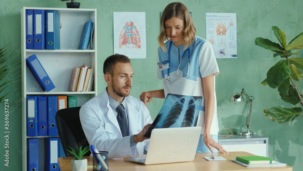 Obraz premium Serious caucasian doctor working at the office on portable laptop. Daring young nurse flirting with therapist trying to seduce him. Sexual harrasment in the hospital. Problems.