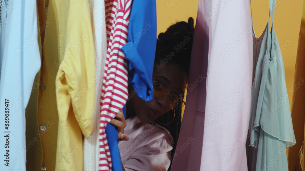 Fototapeta premium Colorful fashion clothing items hanging on store hanger. Young african attractive woman with credit hard goes through clothes selecting from rack. Vip customer.