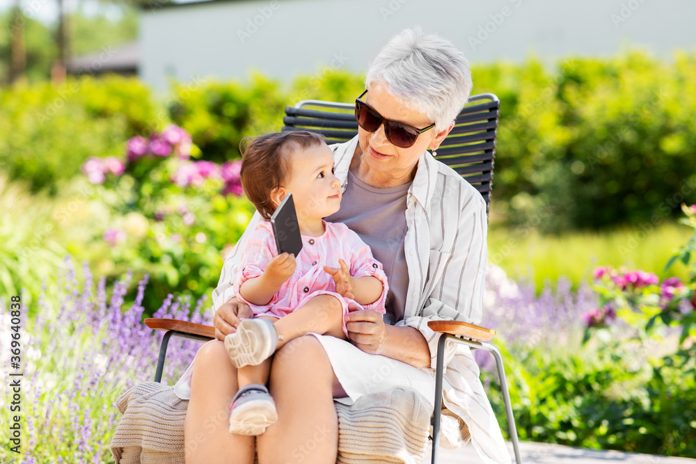 Naklejka premium family, generation and people concept - happy grandmother and baby granddaughter with smartphone at summer garden