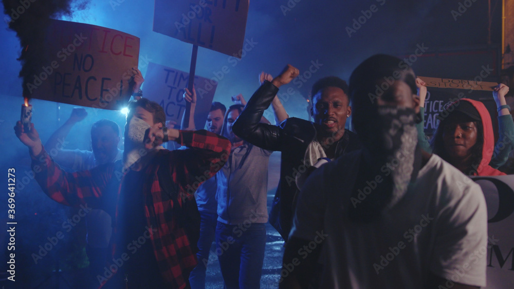 Striking crowd of multi-ethnic men and woman walking on street protest against police brutality with smoke gas grenades at night riot. Rebellion. Antiracism.