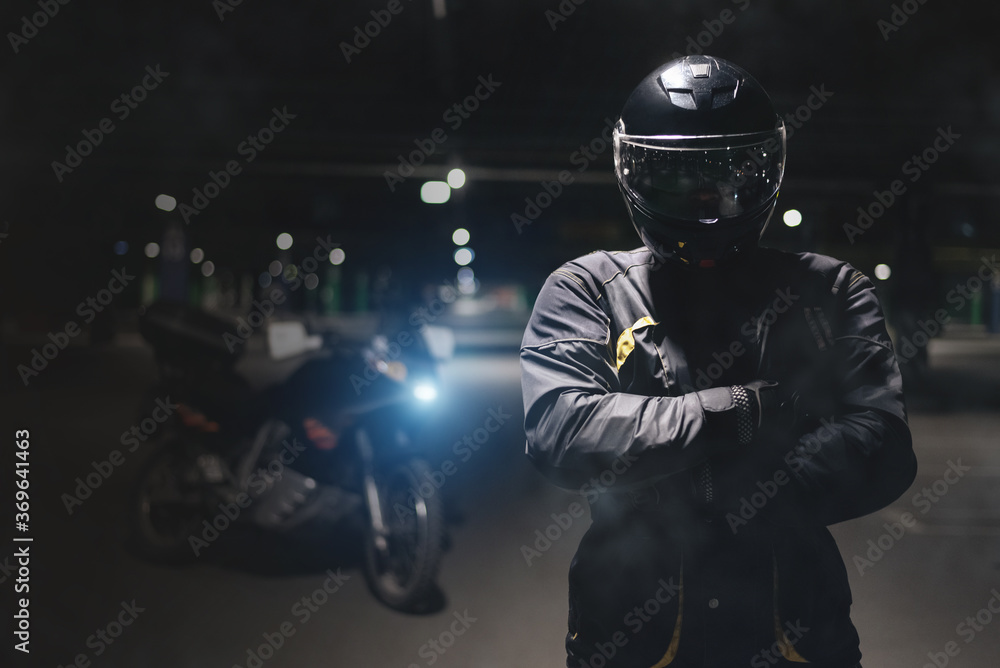 A motorbiker is standing with crossed hands on a dark parking.