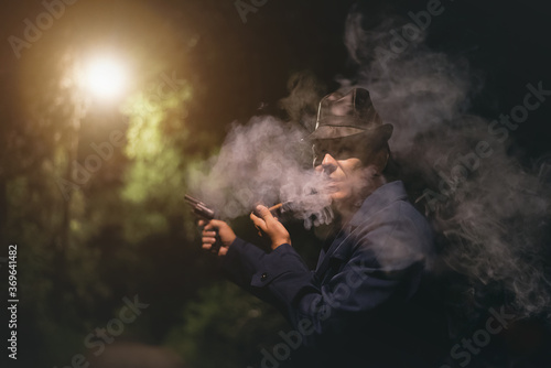 Detective agent in a black leather hat with a gun in hand is smoking a smoming pipe in night park.