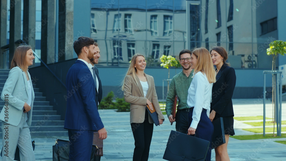 Fototapeta premium Successful corporate team of young workers meeting before work near office building. Cheerful co-workers and their female boss talking on office break outdoor.