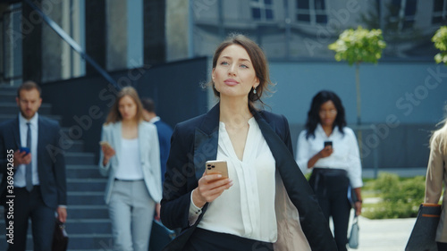 Appealing young caucasian businesswoman with smartphone walks in corporate neighbourhood outdoors. Business people lifestyle. Social distancin Technology. photo