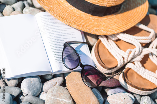 Beach hat on opened book with sunscreen and shoes on pebble beach photo