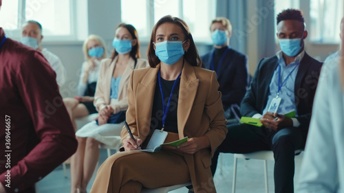 Female journalist in medical mask asking the speaker on business seminar. Diverse corporate people employees clapping hands enjoying start-up presentation. Workspace on quarantine.