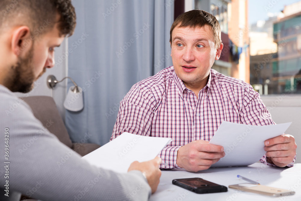 Obraz premium Focused man with male partner attentively reading papers at table in home interior