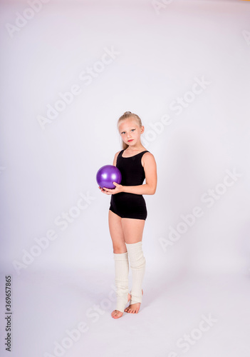 a beautiful gymnast girl in a black swimsuit and white leggings stands with a ball on a white isolated background with space for text