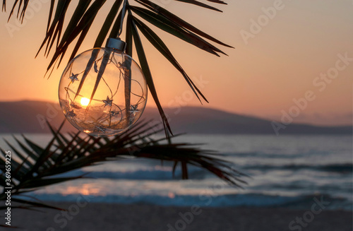 Christmas decorations glass ball during sunset on palm twig over the sea. Ocean beach - Christmas and New Year holidays in hot countries concept. Congratulations, card. Copy space
