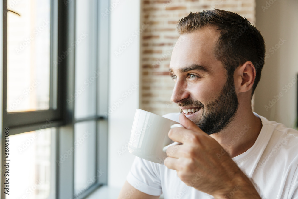 Image of man drinking coffee and smiling while standing near window Image of man drinking coffee and smiling while standing near window
