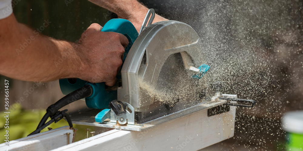 Hampshire, England, UK. 2020. Carpenter holding a circular saw to cut a plank of wood which creates a fine wood dust. Hampshire, England, UK. 2020. Carpenter holding a circular saw to cut a plank of wood which creates a fine wood dust.
