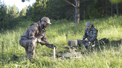Man in camouflage clothes is chopping wood with axe and throwing it into bonfire. Tourist concept. Hunter checks or cleans hunting rifle. Best friends spend leisure weekend in forest nature background