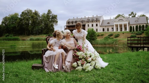 People. Women of different ages are sitting on the couch in the yard. They smile and laugh. They are watching a family album. One girl is a bride. Behind them is an estate with a lake