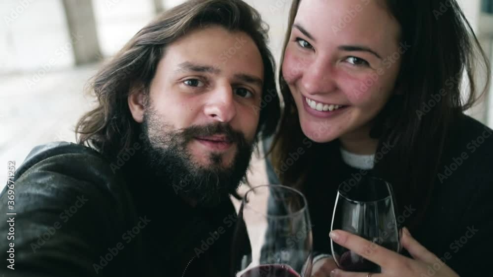 Happy young couple taking video selfie while sitting at table in cafe, drinking and toasting wine, holding wineglasses, laughing and speaking at camera. Front view, closeup. Love and dating concept Happy young couple taking video selfie while sitting at table in cafe, drinking and toasting wine, holding wineglasses, laughing and speaking at camera. Front view, closeup. Love and dating concept