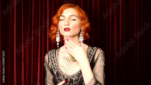 Retro woman with pale skin and red hair in black vintage dress dance against red background in theater. Vintage redhead woman with red lips, blue eyes and retro hairstyle. Beautiful silver earrings