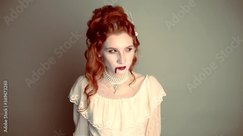 Young vampire woman with blood on face. Scary creepy character. Pale skin demon in white dress on gray background. Redhead model with red lips. Outfit for halloween. Vampire with open mouth and fangs