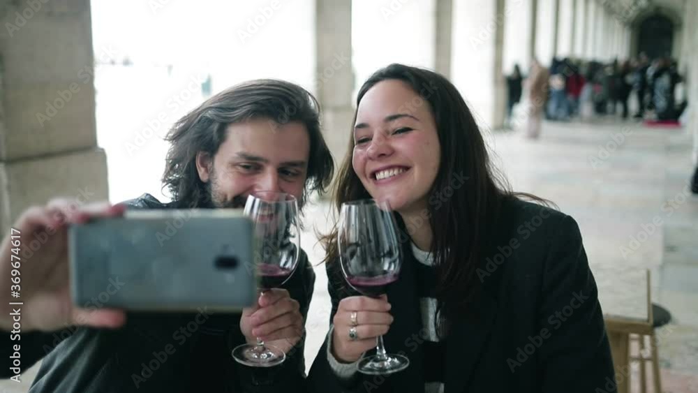 Happy young couple having video call on phone while sitting at table in cafe, drinking wine, toasting, laughing and speaking at screen. Front view, closeup. Communication concept Happy young couple having video call on phone while sitting at table in cafe, drinking wine, toasting, laughing and speaking at screen. Front view, closeup. Communication concept