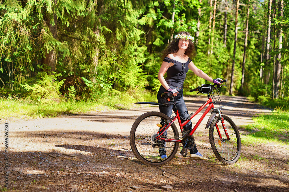 Happy woman in wreath of daisies with bike alone in the bright green forest