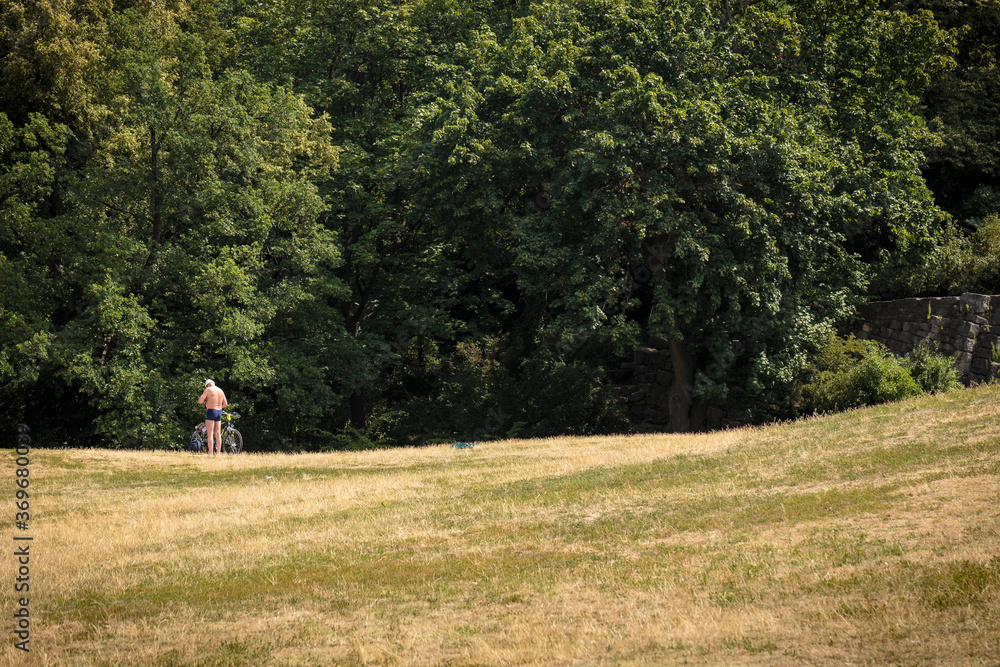 Wide angle view of 60 year old male sunbather standing shirtless next to bicycle on the ample lawn of the Wide angle view of 60 year old male sunbather standing shirtless next to bicycle on the ample lawn of the