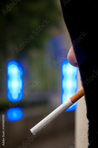 Vertical view of mysterious man with hidden face covered by black hoodie. Unrecognisable man with unlit cigarette hanging from mouth. In the background, bokeh from unsharp blue neon light