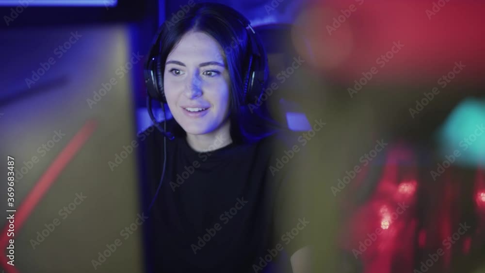 Beautiful young woman sits in headphones and plays an online game in a computer club. Woman enthusiastically plays and talks on the microphone with her partners. Portrait view. Beautiful young woman sits in headphones and plays an online game in a computer club. Woman enthusiastically plays and talks on the microphone with her partners. Portrait view.