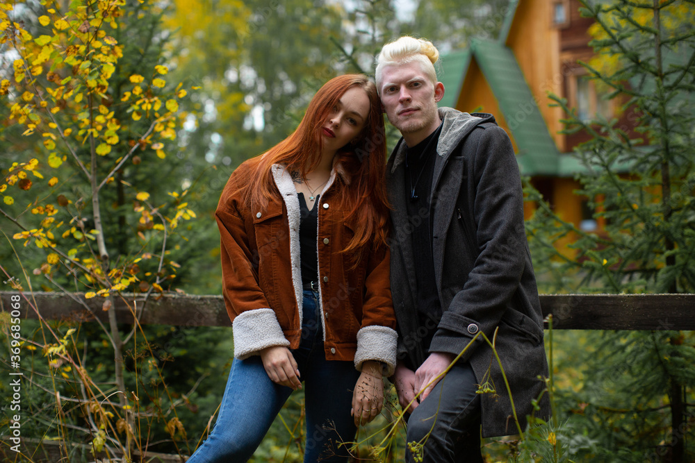 Fototapeta premium A cute couple of young guys - a red-haired girl and an albino guy, stand huddled together at the fence against the background of the forest and a wooden house with a green roof
