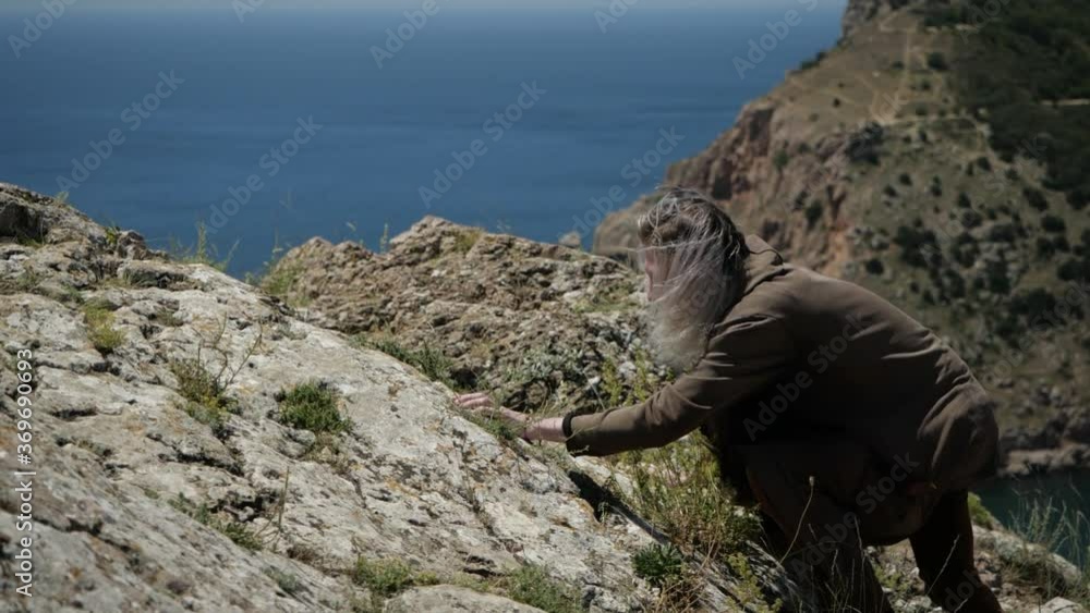 A young man in brown clothes hardly climbs up the rock, reaching the top he stands up in full height and looks at the seascape opened in front of him A young man in brown clothes hardly climbs up the rock, reaching the top he stands up in full height and looks at the seascape opened in front of him