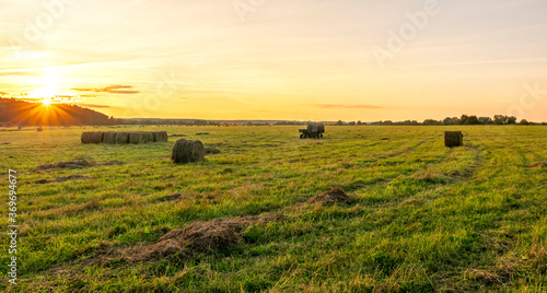 Scenic view at beautiful sunset in green shiny field with hay stacks  bright cloudy sky  country road and golden sun rays with glow  summer valley landscape