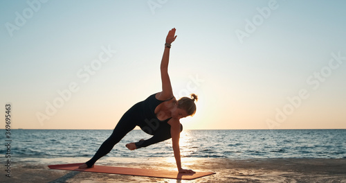 The woman balanced her body in a yoga pose with an emphasis on her left hand against the background of the sea, blocking the sun. Human harmony with marine nature.