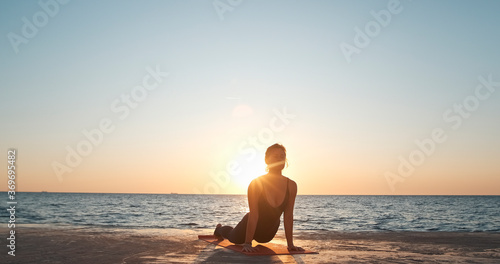 A girl performs an exercise from yoga, doing a plank with an emphasis on her arms and arching her back against the background of the sea in the early morning.
