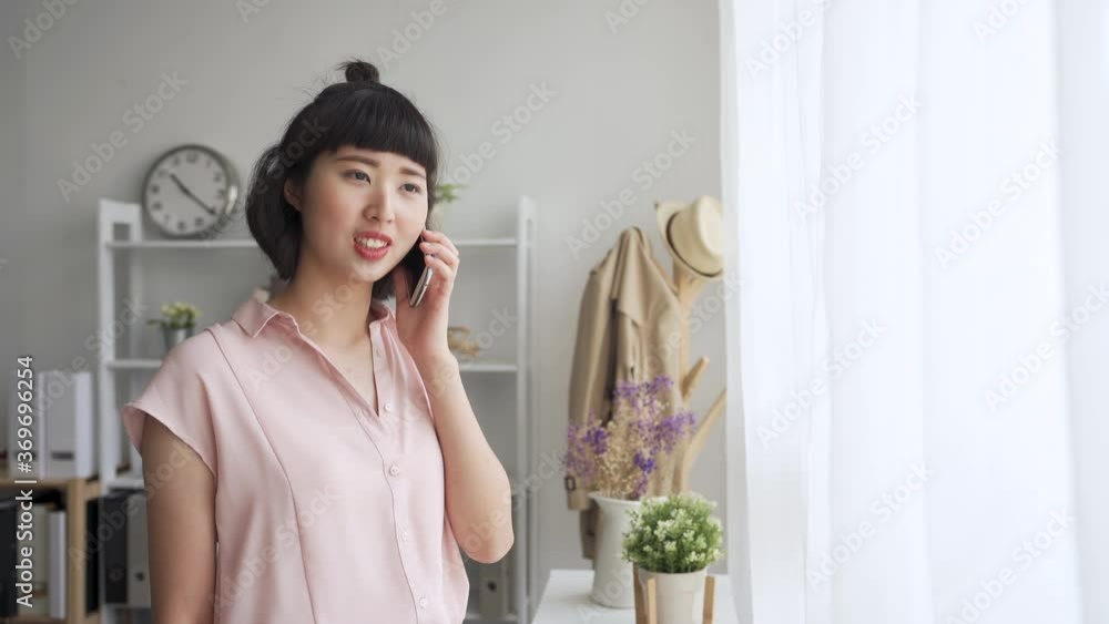 attractive young asian chinese woman talking on mobile phone call with friend in home living room. lady standing by window and chatting on cellphone in house. smiling female relax gossip online. attractive young asian chinese woman talking on mobile phone call with friend in home living room. lady standing by window and chatting on cellphone in house. smiling female relax gossip online.
