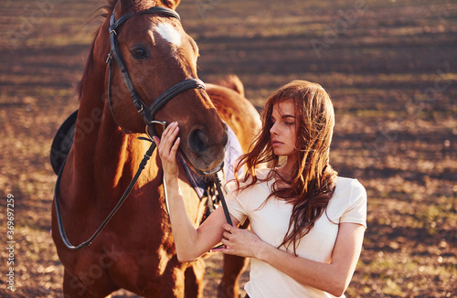 Young woman standing with her horse in agriculture field at sunny daytime