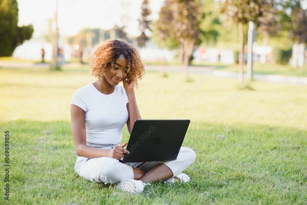Young attractive dark-skinned college student wearing tank top and jeans sitting on the lawn at campus on sunny day, working on her thesis using laptop computer, looking busy and concentrated Young attractive dark-skinned college student wearing tank top and jeans sitting on the lawn at campus on sunny day, working on her thesis using laptop computer, looking busy and concentrated