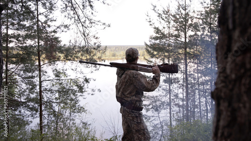 View from back of hunter in camouflage puts hunting rifle on his shoulder and looks at river from forest. Outdoor activity concept. Summer hiking in wilderness area.