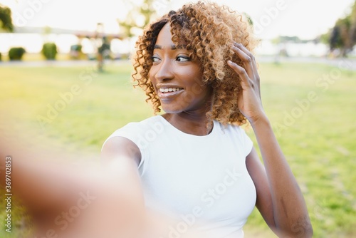 Closeup portrait of beautiful african american woman sitting outdoors in the park taking selfie and messaging via social networks using smart phone using free wireless. People and lifestyle concept