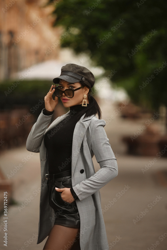 Adult fashionable model in a trendy trench coat, leather shorts and hat looking in the camera and posing outdoors