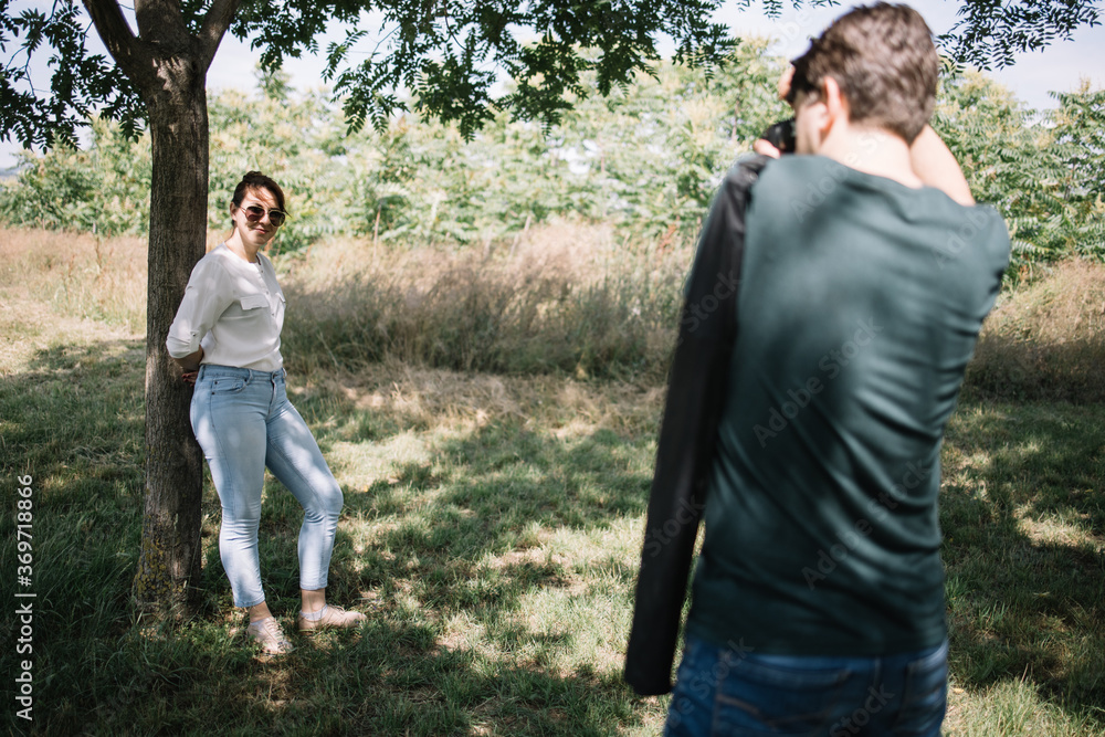Photographer taking photo session of female model in forest. Lensman taking photos of pretty woman who is leaning her back against tree in park. Photographer taking photo session of female model in forest. Lensman taking photos of pretty woman who is leaning her back against tree in park.