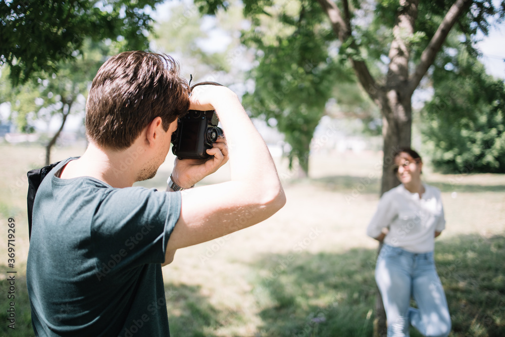 Man taking photos of girl in park. Back view of man using DSLR camera while taking photos of out of focus girl who is posing near tree in nature. Man taking photos of girl in park. Back view of man using DSLR camera while taking photos of out of focus girl who is posing near tree in nature.