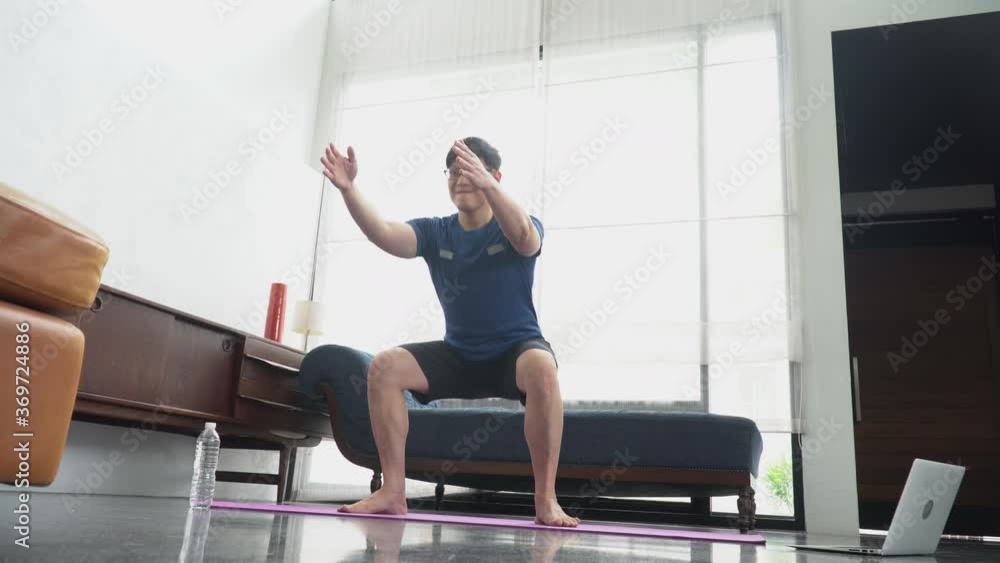 young asian fit man in sportswear doing squat exercises While watching video training fitness online tutorials on laptop in living room at home . sport male workout in quarantine young asian fit man in sportswear doing squat exercises While watching video training fitness online tutorials on laptop in living room at home . sport male workout in quarantine